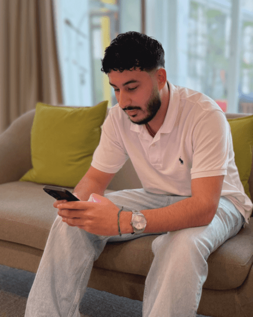Mann sitzt auf einem Sofa und schaut auf sein Smartphone. Im Hintergrund sind helle Fenster und grüne Kissen zu sehen.