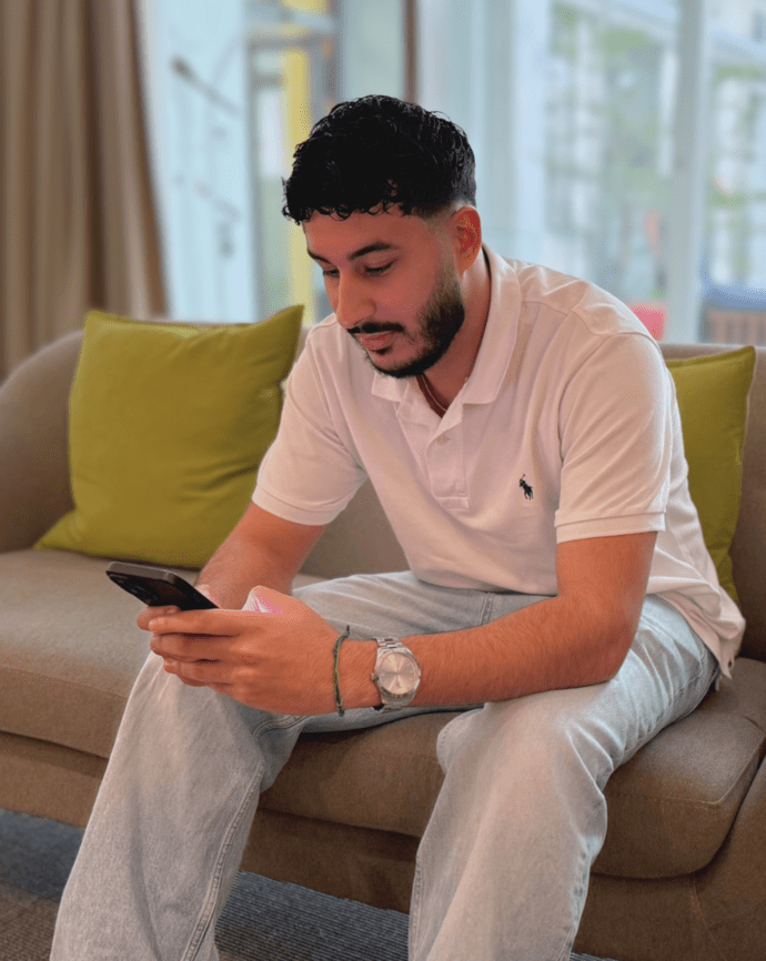 Mann sitzt auf einem Sofa und schaut auf sein Smartphone. Im Hintergrund sind helle Fenster und grüne Kissen zu sehen.