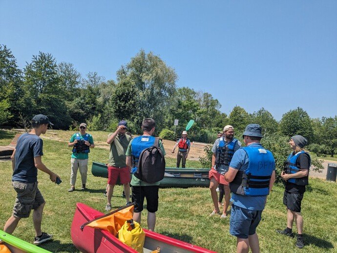 Personen mit Schwimmwesten stehen neben Kanus auf einer Wiese, umgeben von Bäumen, unter blauem Himmel.