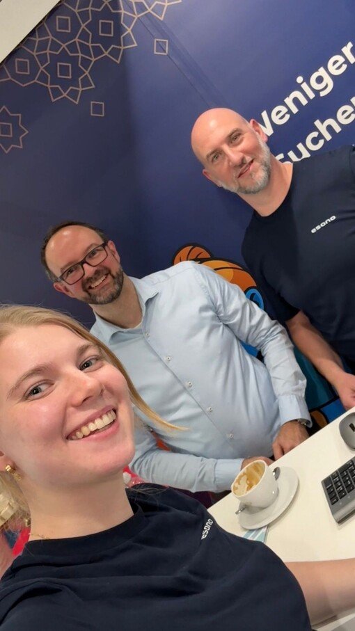 Drei Personen lächeln in die Kamera, stehend vor einem blauen Hintergrund mit geometrischen Mustern, Kaffee auf dem Tisch.