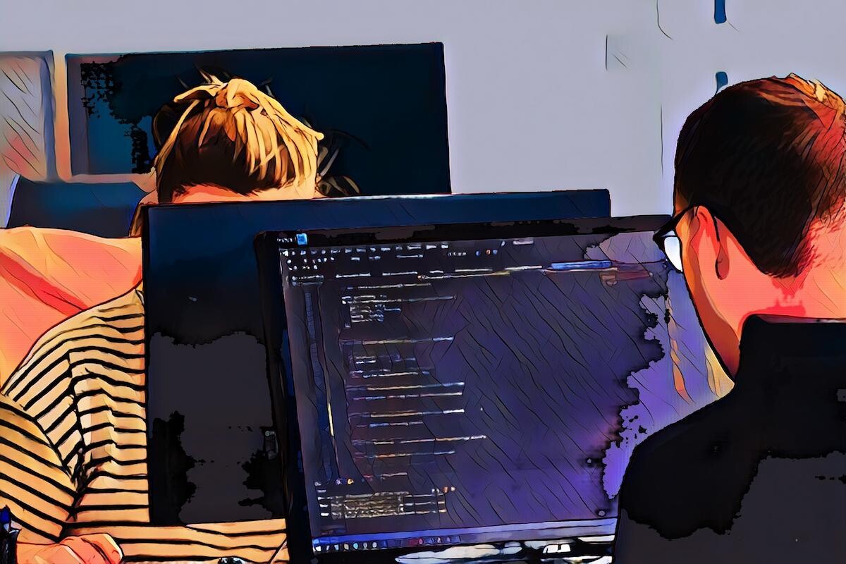 Zwei Personen arbeiten an Computern, einer zeigt Code auf dem Bildschirm.