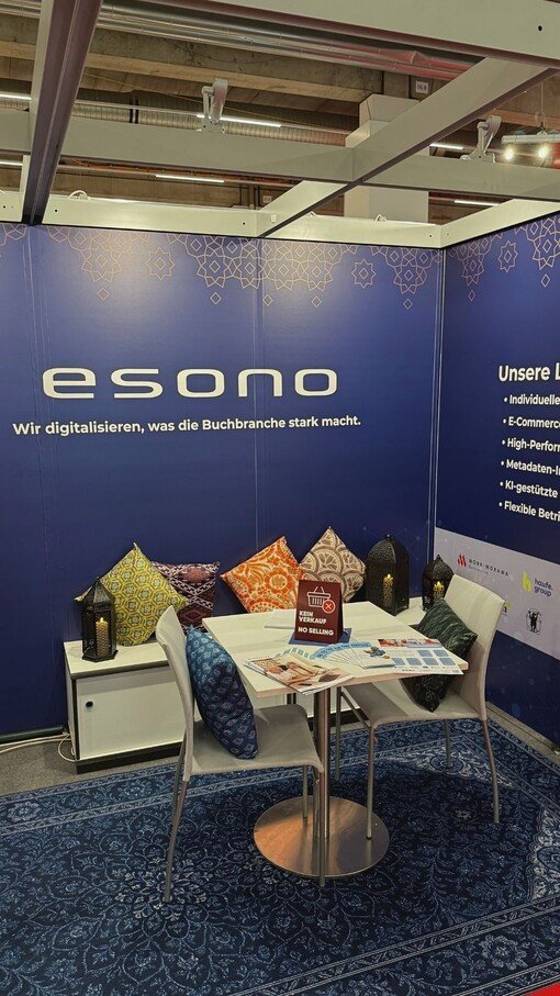 Messestand mit blauem Hintergrund, Tisch, zwei Stühlen, bunten Kissen und Prospekten. Text: "esono - Wir digitalisieren, was die Buchbranche stark macht."