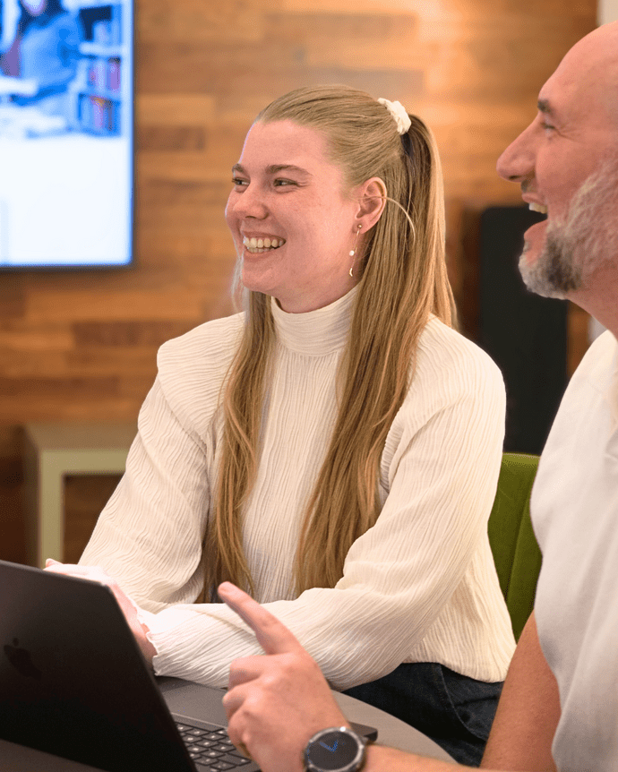 Zwei Personen lächeln in einem Besprechungsraum. Sie sitzen vor einem Laptop. Der Raum hat eine hölzerne Wandverkleidung.