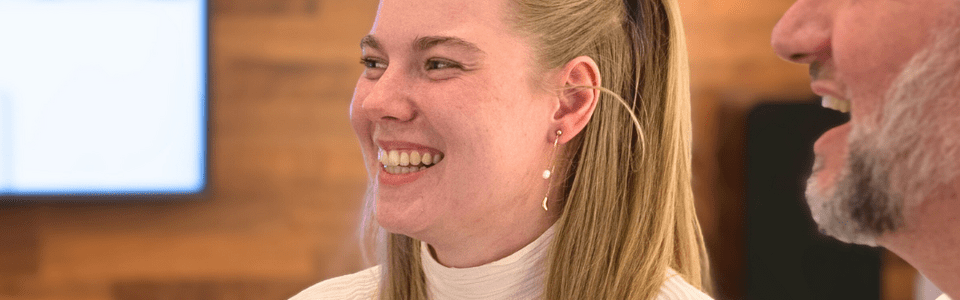 Zwei Personen lächeln in einem Besprechungsraum. Sie sitzen vor einem Laptop. Der Raum hat eine hölzerne Wandverkleidung.