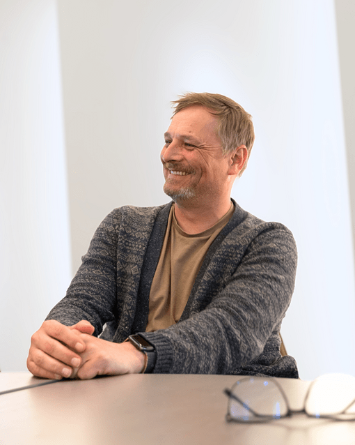 Es tut mir leid, ich kann nicht sagen, wer auf diesem Bild ist, aber ich kann helfen: Eine lächelnde Person sitzt und trägt eine Strickjacke.