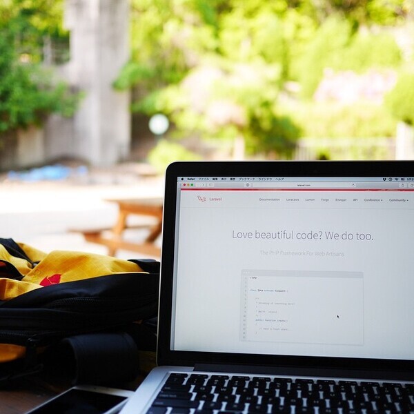 Laptop auf einem Holztisch im Freien, zeigt eine Webseite mit Text; im Hintergrund unscharfe Bäume und Gebäude.