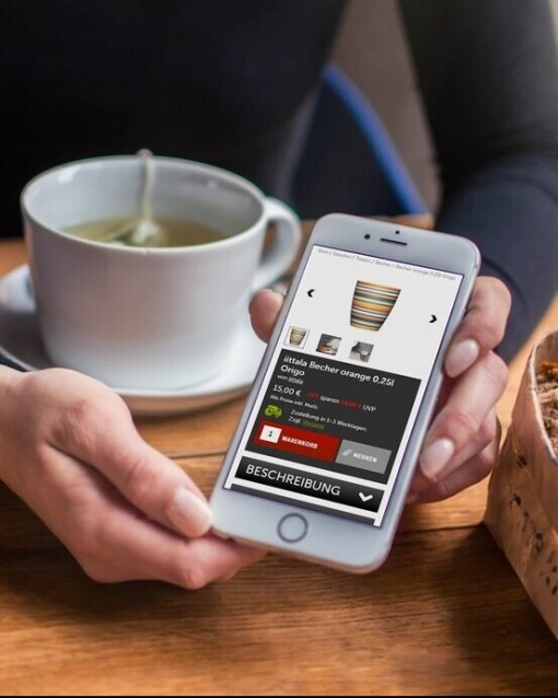 Person hält Smartphone mit Onlineshop; im Hintergrund eine Tasse Tee und ein Brotkorb auf Holztisch.