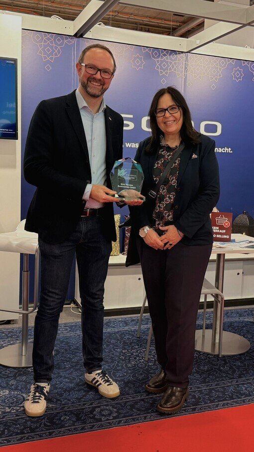 Personen halten eine Trophäe an einem Messestand. Hintergrund mit blauem Design und Textausschnitt.