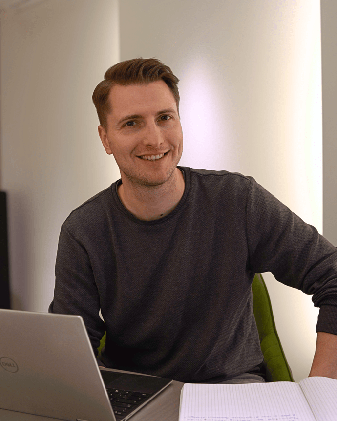 Eine Person sitzt an einem Tisch mit Laptop und Notizbuch, lächelt in die Kamera.