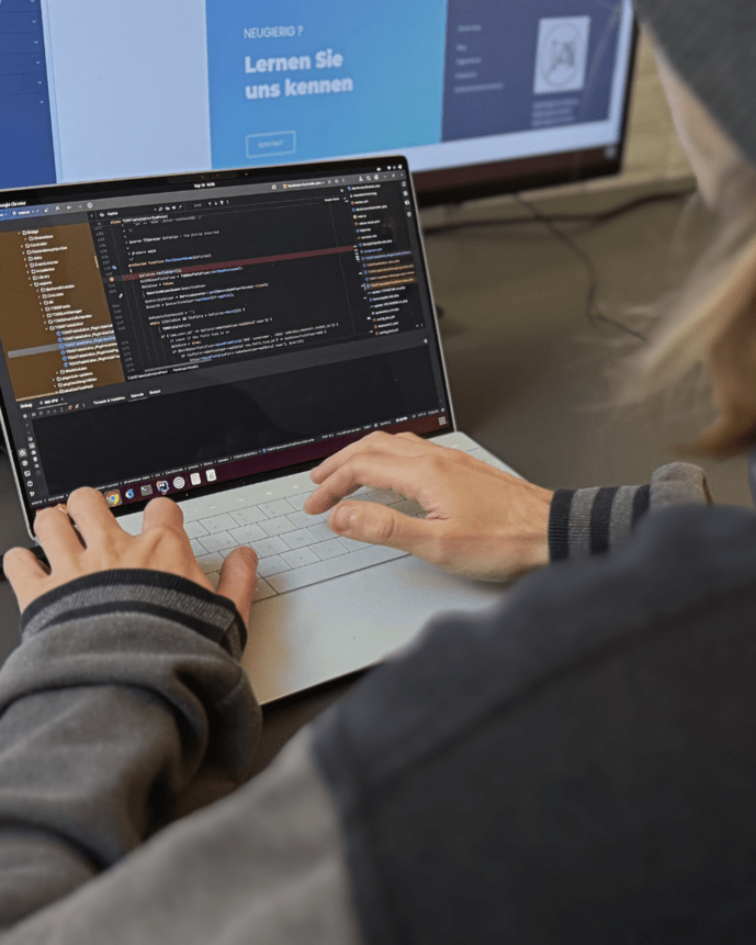 Person arbeitet an einem Laptop mit geöffnetem Code-Editor, im Hintergrund zwei Monitore.