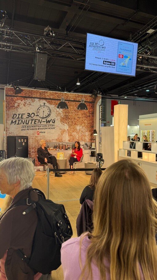 Bühne mit Interview, roter Stuhl, Zuschauer im Vordergrund, Backsteinwand mit Logo "Die 30 Minuten-WG", Messeatmosphäre.