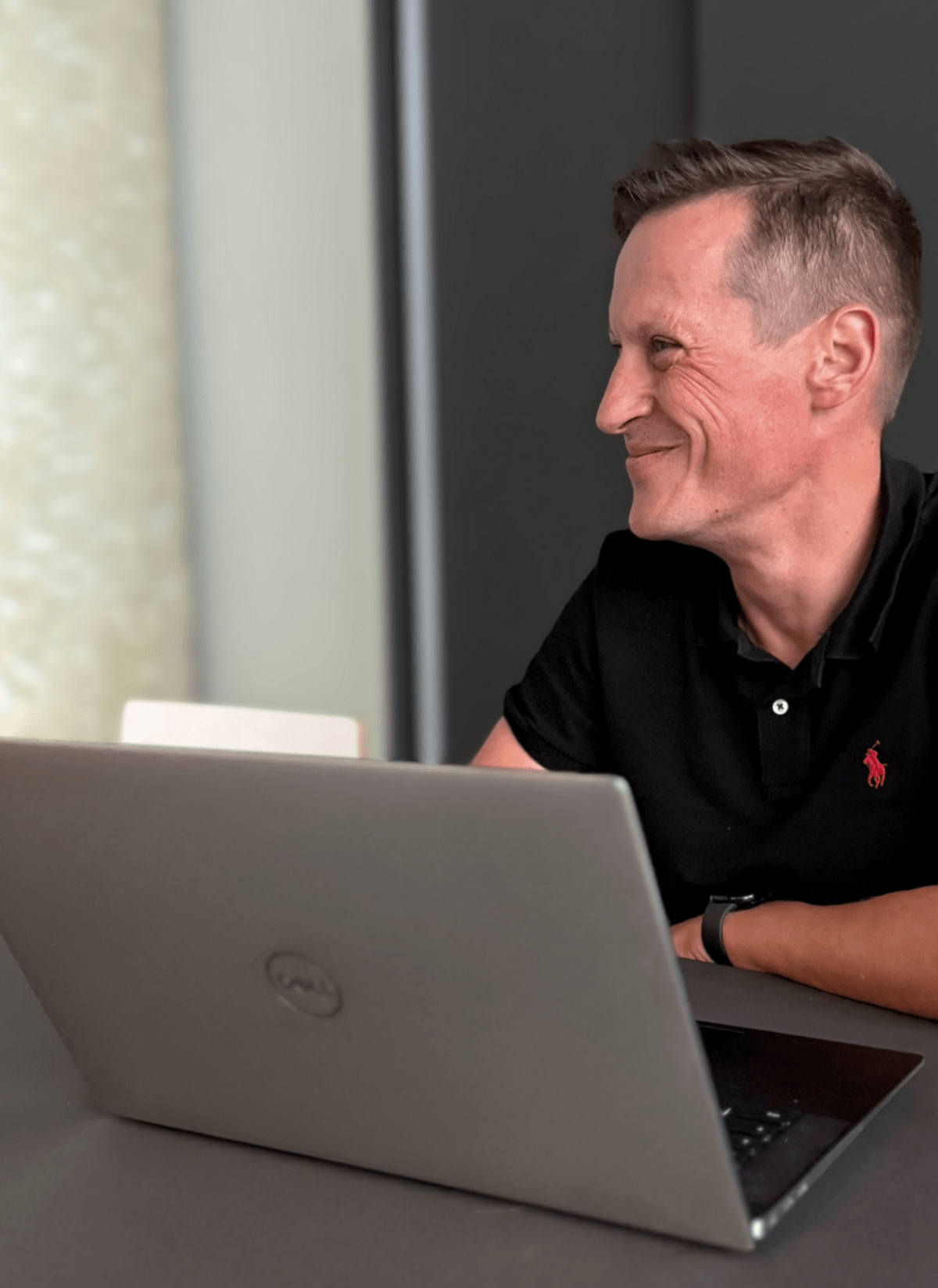 Eine Person sitzt lächelnd an einem Tisch mit einem geöffneten Laptop in einem Büro.