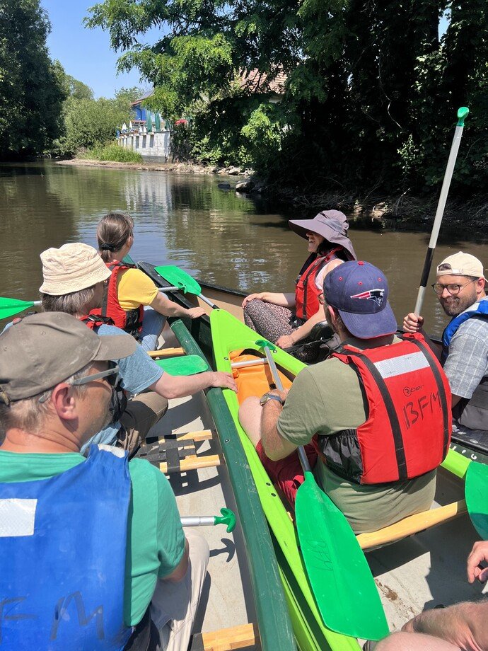 Gruppe von Menschen in Kanus mit Schwimmwesten auf einem Fluss, umgeben von Bäumen bei sonnigem Wetter.