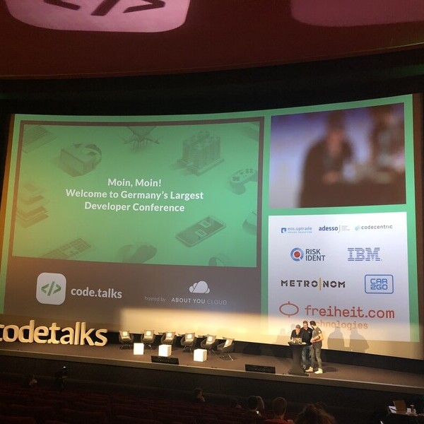 Große Leinwand auf einer Entwicklerkonferenzbühne mit mehrsprachiger Begrüßungsnachricht und mehreren sprechenden Personen.