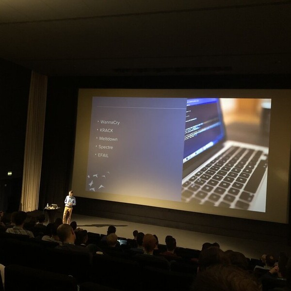 Person hält Vortrag vor Publikum mit großer Leinwand und Laptop, Bildschirm zeigt Code und Textinhalte.