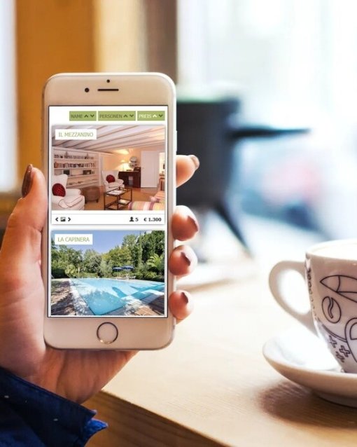 Smartphone zeigt Ferienunterkünfte, gehalten von Person in Café, daneben eine dekorierte Tasse.