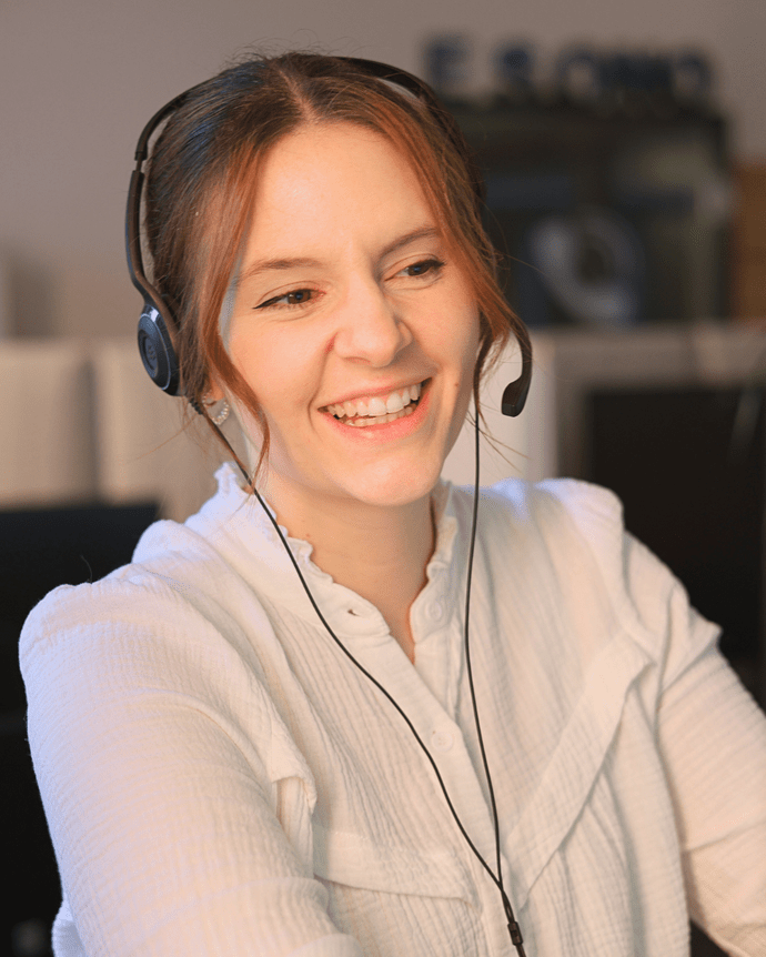 Eine Frau mit Headset lacht und arbeitet am Computer in einem Büro.