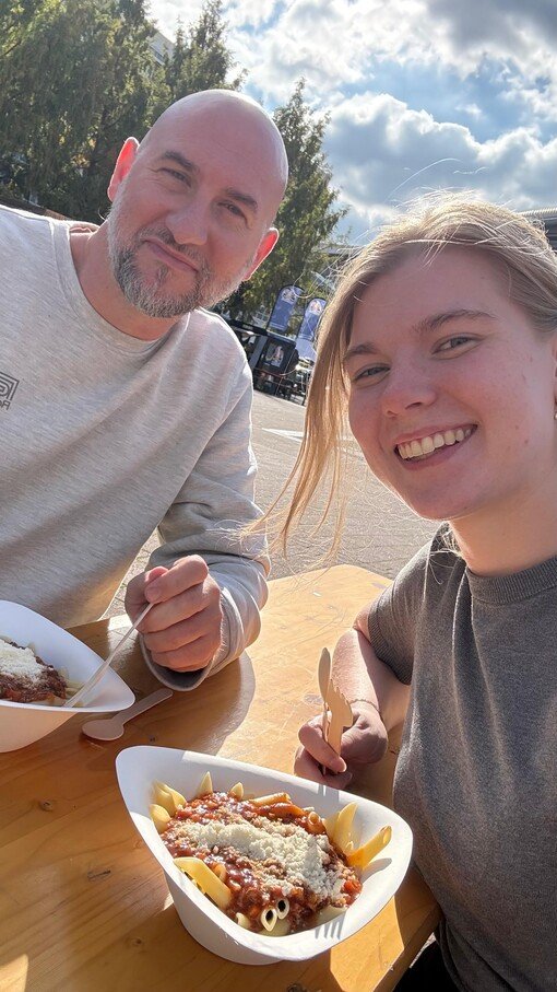 Zwei Personen essen draußen Pasta an einem Holztisch. Es ist sonnig, Bäume im Hintergrund.