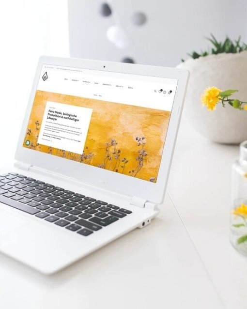 Laptop auf weißem Schreibtisch mit Blumen und Kaffeebecher, zeigt eine Website mit gelbem Hintergrund.