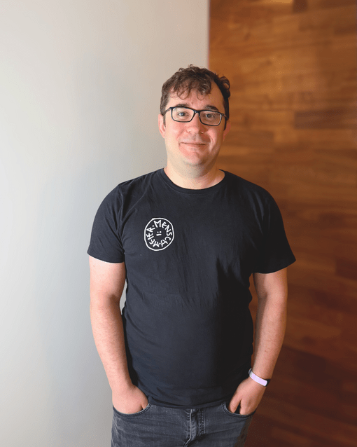 Ein Mann mit Brille trägt ein schwarzes T-Shirt vor einer teils weißen, teils holzverkleideten Wand.