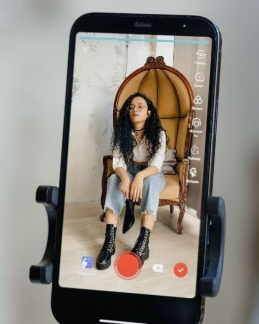 Smartphone im Stativ zeigt Frau in Jeans und Stiefeln auf dem Bildschirm, die auf einem Sessel sitzt.