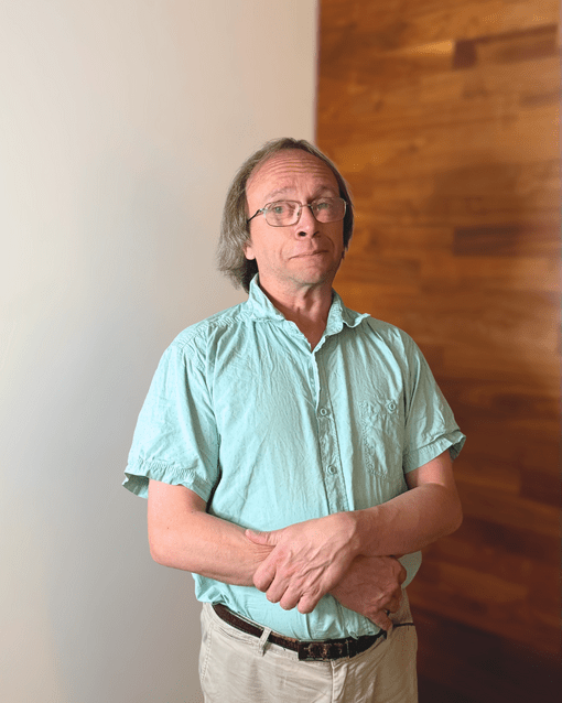 Eine Person mit Brille und hellgrünem Hemd steht vor einer Holzwand.