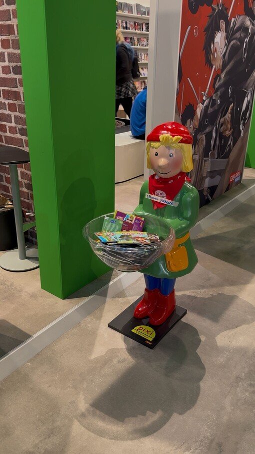 Kinderfigur als Werbedisplay mit Korb voller Prospekte in einer Verkaufsausstellung, umgeben von grünen Wänden.
