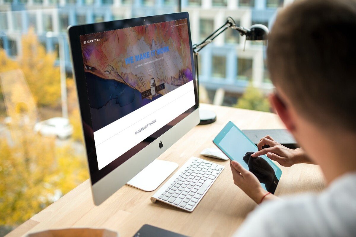 Person nutzt Tablet am Schreibtisch mit iMac und Tastatur; Fokus auf digitaler Interaktion in moderner Büroumgebung.