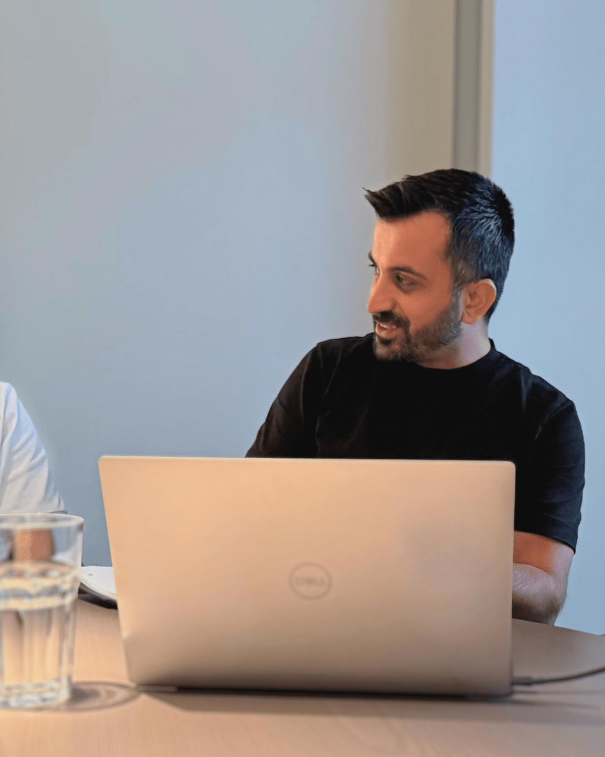 Zwei Männer unterhalten sich am Tisch; Laptop und Glas Wasser sichtbar.