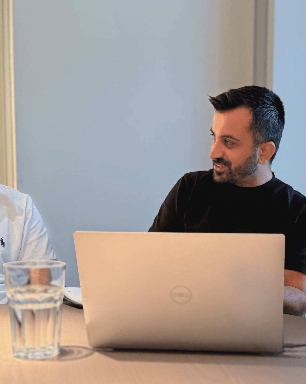 Zwei Männer unterhalten sich am Tisch; Laptop und Glas Wasser sichtbar.