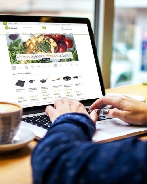 Person tippt auf Laptop in Café, Webseite mit Küchengeräten geöffnet; daneben Kaffee und Limonade mit Minze.