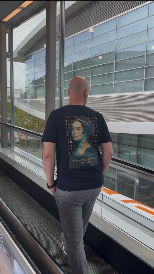 Person mit schwarzem T-Shirt und Ada-Lovelace-Motiv auf Rolltreppe vor modernem Gebäude.