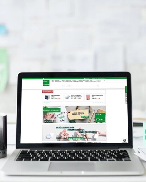 Laptop auf Schreibtisch vor weißer Wand, geöffnet mit Webseite auf dem Bildschirm; daneben Tasse und Dokumente.