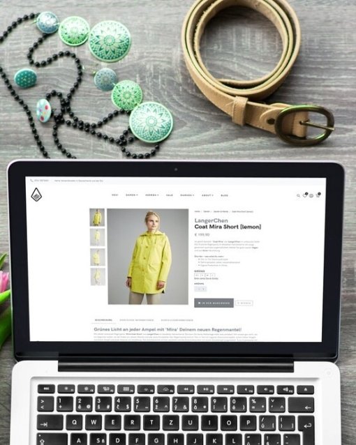 Laptop mit Online-Shop-Seite, umgeben von rosa Tulpen, Kaffee, Accessoires und Kleidung auf Holzuntergrund.