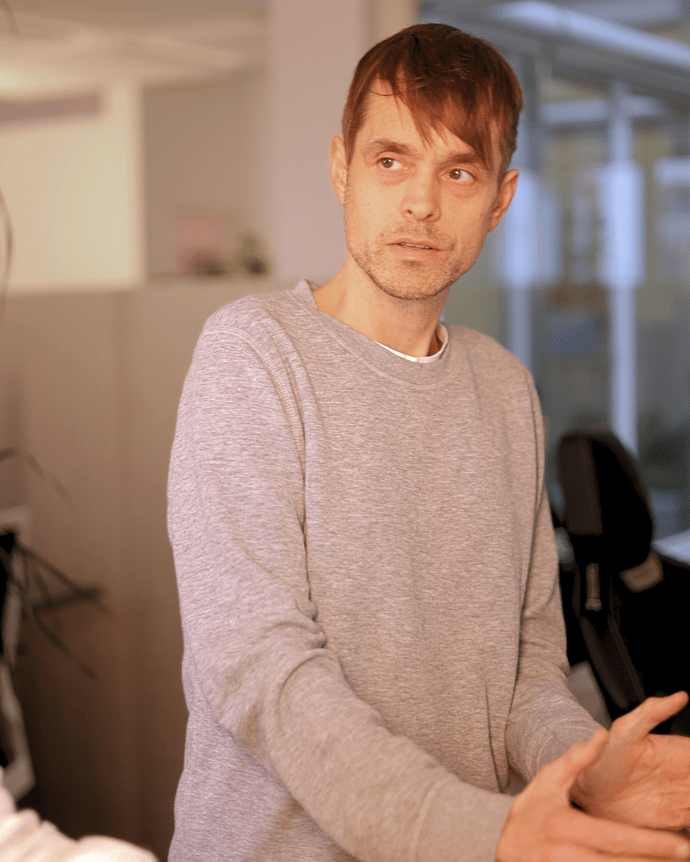 Eine Person in einem Büro spricht gestikulierend, trägt einen grauen Pullover. Hintergrund ist verschwommen, zeigt Büromöbel.