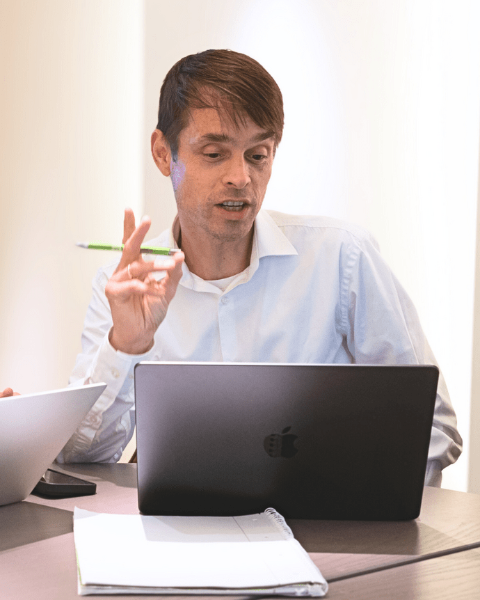 Eine Person konzentriert sich auf einen Laptop am Tisch, hält einen Stift und scheint zu sprechen.