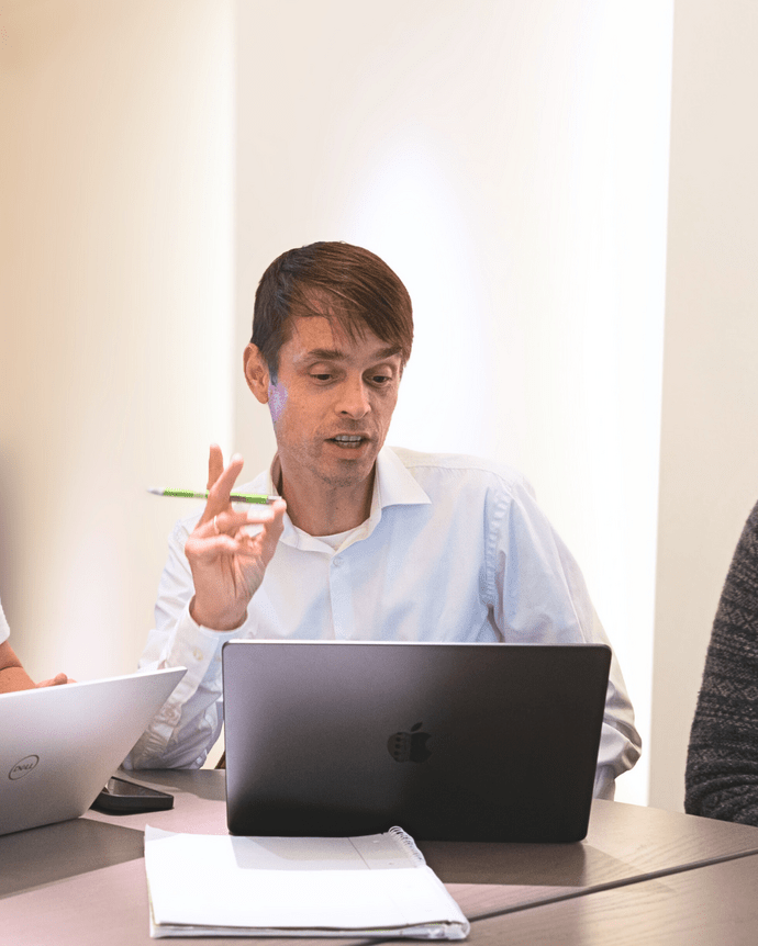 Eine Person konzentriert sich auf einen Laptop am Tisch, hält einen Stift und scheint zu sprechen.