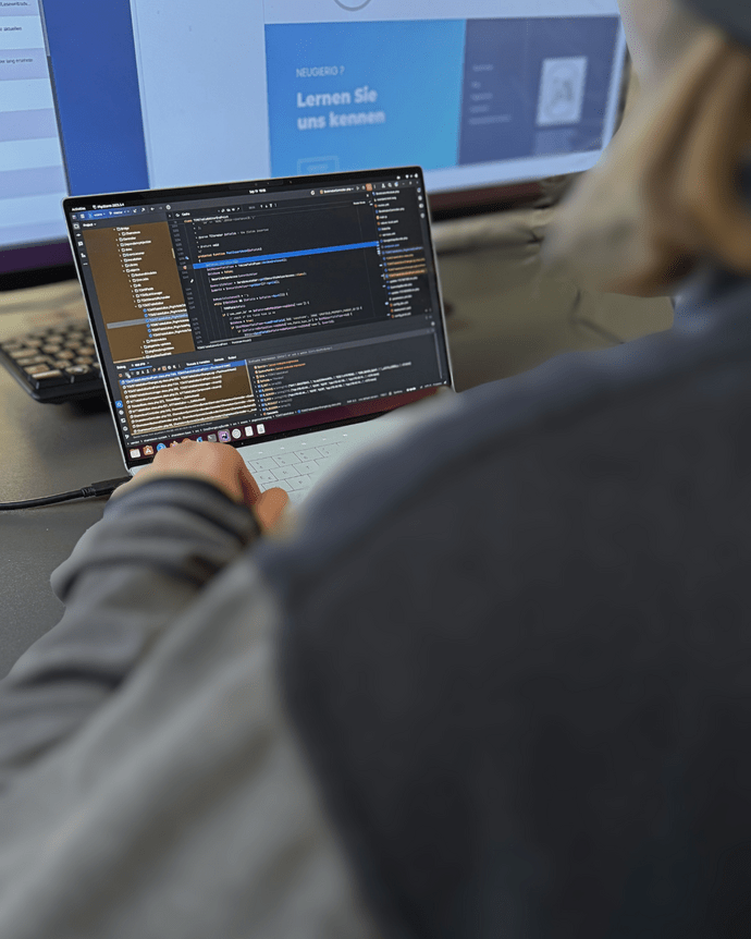 Person arbeitet an einem Laptop mit geöffnetem Code-Editor; im Hintergrund unscharfer Bildschirm.