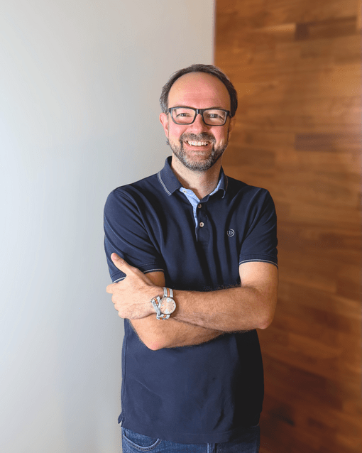 Person mit Brille und Bart steht lächelnd mit verschränkten Armen vor einer Holzwand. Trägt ein dunkles Polohemd und eine Armbanduhr.