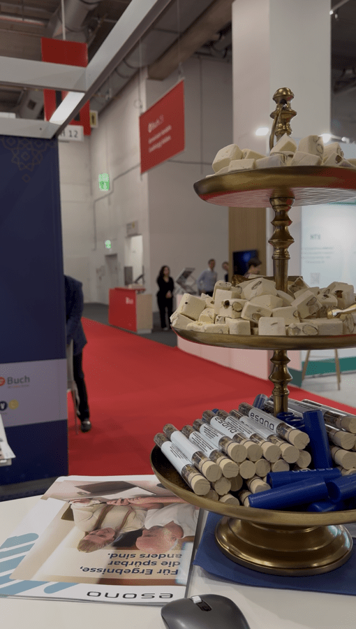 Messestand mit zweistöckiger Etagere, bestückt mit Werbeartikeln; im Hintergrund sind Messebesucher und -schilder erkennbar.