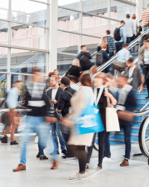 Menschenmenge in Bewegung auf einer Messe, einige auf Rolltreppen, moderne Architektur im Hintergrund.
