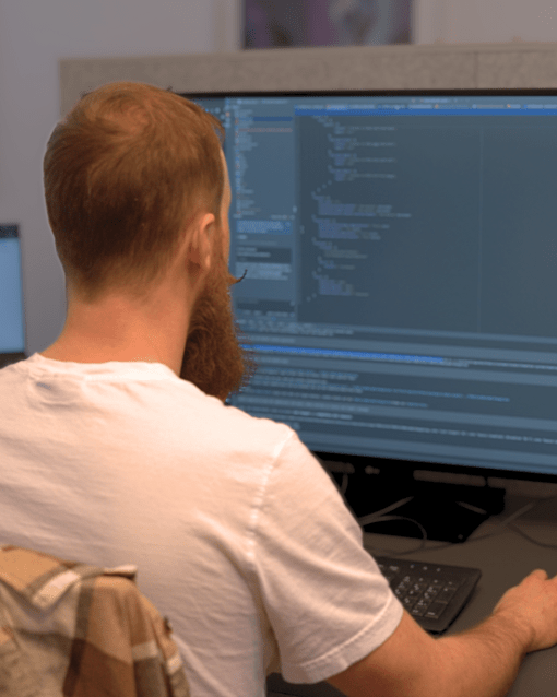 Mann arbeitet an einem Computer mit großem Monitor, Programmiercode auf dem Bildschirm sichtbar.