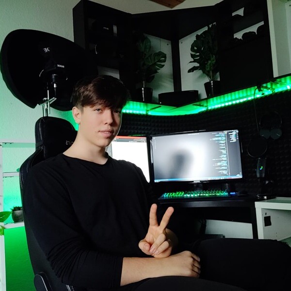 Person sitzend vor einem Computer mit mehreren Bildschirmen und grüner Beleuchtung, zeigt ein Peace-Zeichen.